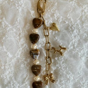 Peut inclure: Un porte-clés doré avec un mousqueton, une chaîne et des breloques décoratives. Les breloques comprennent des perles en forme de cœur marron, des fausses perles, un cœur, un nœud et un ballon en forme de chien.