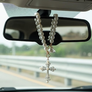 May include: A car charm hanging from a rearview mirror. The charm features a strand of white faux pearls, a gold-colored accent, and a silver-colored cross pendant adorned with clear crystals and pearls. The cross is a religious symbol.