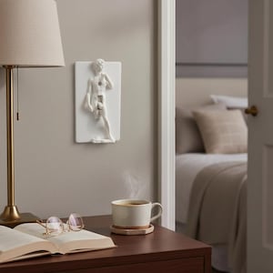May include: A white decorative light switch cover featuring a sculpted figure of David. The switch is in the center of the figure. The cover is mounted on a light gray wall. A lamp, book, and coffee cup are on a nearby table.
