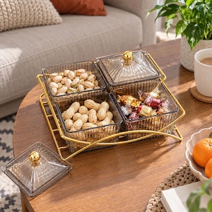 May include: A set of four square, ribbed, dark-tinted glass snack containers arranged in a gold-toned metal wire rack. Each container features a matching glass lid with a small gold spherical knob. The containers hold pistachios, peanuts, and candies.