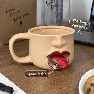 Puede incluir: Una taza de cerámica beige con forma de rostro humano con nariz y labios. La boca está abierta, revelando una lengua roja unida a un resorte metálico en el interior. El texto en la imagen dice The tongue can move! y Spring inside.