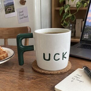 Könnte beinhalten: Ein weißer Keramikbecher mit einem dunkelgrünen Henkel in Form des Buchstabens F. Der Becher trägt den dunkelgrünen Schriftzug UCK auf der Vorderseite. Er steht auf einem runden Korkuntersetzer, enthält eine dunkle Flüssigkeit und steht auf einer Holzoberfläche.