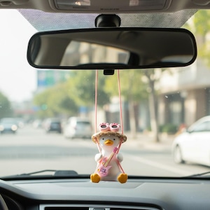 May include: A car accessory featuring a white penguin figurine wearing sunglasses and a straw hat, holding a pink guitar. The penguin is suspended by pink strings from the rearview mirror. The background shows a street scene.