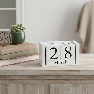 May include: A white wooden perpetual calendar displays "28 March" in black block letters. A small green potted succulent and a stack of books sit nearby on a light-colored wooden surface. A gold geometric decoration is also visible.