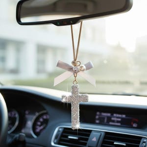 May include: A car charm featuring a sparkling, silver cross pendant. The cross is adorned with numerous small, reflective embellishments. It hangs from a light-colored cord, accented with a gray bow and pearl-like beads. The charm is suspended in front of a car's dashboard and rearview mirror.