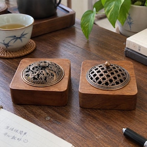 May include: Two square wooden incense burners with rounded corners. Each features a circular metal lid with intricate cut-out designs, one displaying a dragon motif and the other a woven lattice pattern, both set into the smooth brown wood base.