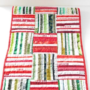 May include: Red, green, and white striped fabric table runner with a quilted design. The fabric is made of various strips of fabric sewn together. The runner is approximately 16 inches wide and 36 inches long.