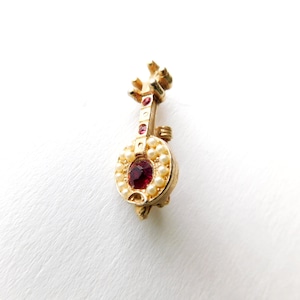 May include: A gold-tone brooch in the shape of a lute with a red rhinestone in the center and white pearl accents.
