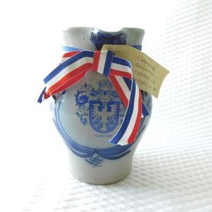 May include: A light blue ceramic pitcher with a blue and white design, a red, white, and blue ribbon, and a tag. The pitcher features a crest with an eagle and the word "Frankfurt". The ribbon is tied around the neck of the pitcher.