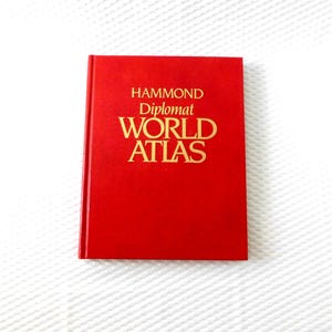 May include: A red hardcover book titled "HAMMOND Diplomat WORLD ATLAS" in gold lettering. The book is closed and centered, with a plain red cover. The background is a white, textured surface.