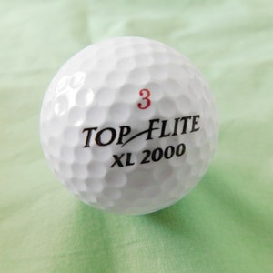 May include: White golf ball with black text that reads "TOP-FLITE XL 2000" and a red number "3" printed on the ball.