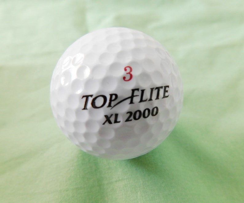 May include: White golf ball with black text that reads "TOP-FLITE XL 2000" and a red number "3" printed on the ball.
