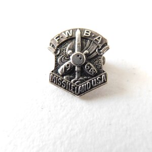 May include: A silver-toned metal pin with a shield shape. The pin features a missile design with the text "FWBA" at the top and "MISSILE LAND USA" at the bottom. The numbers "1968" are also visible.