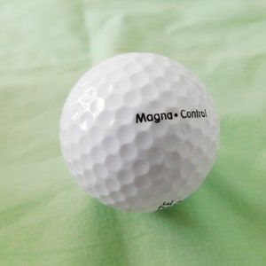 May include: A white golf ball with the text "Magna Control" printed on it.