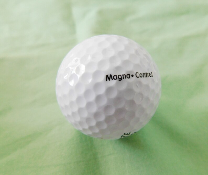 May include: A white golf ball with the text "Magna Control" printed on it.