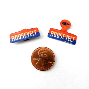 May include: Two vintage enamel pins with the word "ROOSEVELT" in white letters on a blue and red background. The pins are about the size of a US penny.