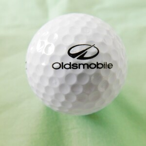 May include: A white golf ball with a black Oldsmobile logo and a stylized arrow design.