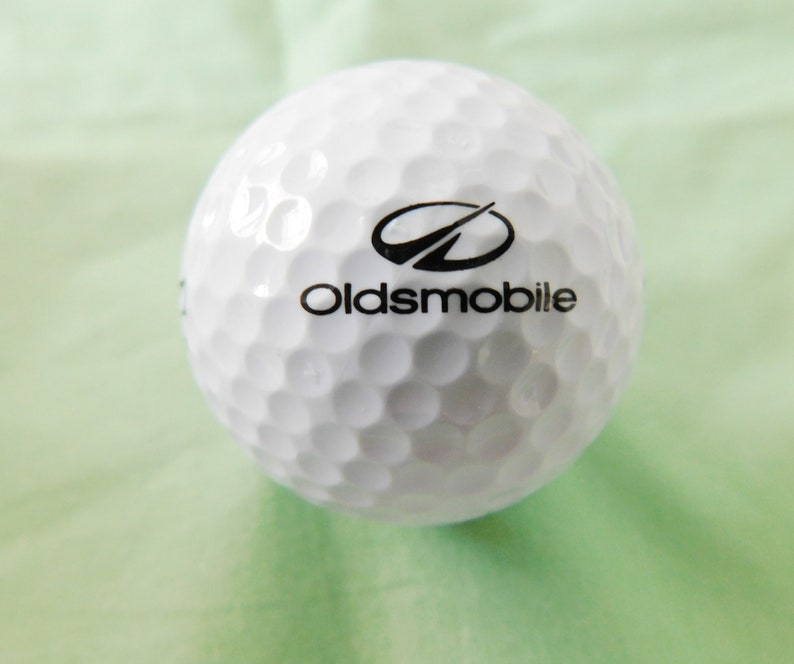 May include: A white golf ball with a black Oldsmobile logo and a stylized arrow design.