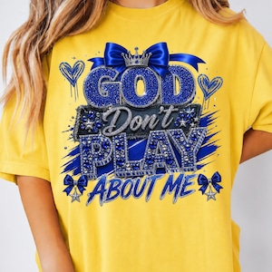 Può includere: Una t-shirt giallo brillante con un design grafico che riporta il testo GOD Don't PLAY ABOUT ME. Il design include scritte blu glitterate, una corona argentata, fiocchi blu, forme a cuore e accenti a stella in una composizione a strati.