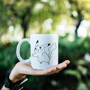May include: White ceramic mug featuring a black outline illustration of Pikachu. The design shows Pikachu with a happy expression and outstretched arm. The mug has a curved handle and is held in a hand.