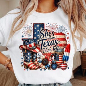 Pode incluir: Uma camisola branca de gola redonda com um gr&aacute;fico do estado do Texas com padr&otilde;es da bandeira americana, fogo de artif&iacute;cio, um chap&eacute;u de cowboy, uma bota, um girassol, um frasco, um gelado e &oacute;culos em forma de cora&ccedil;&atilde;o com o texto She's From Texas I Can Tell.