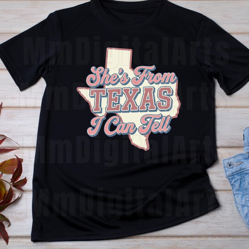 May include: Black t-shirt with a cream-coloured outline of the state of Texas, with the words "She's From TEXAS I Can Tell" in pink and blue. The shirt is made of a soft material. The design is vintage-inspired.
