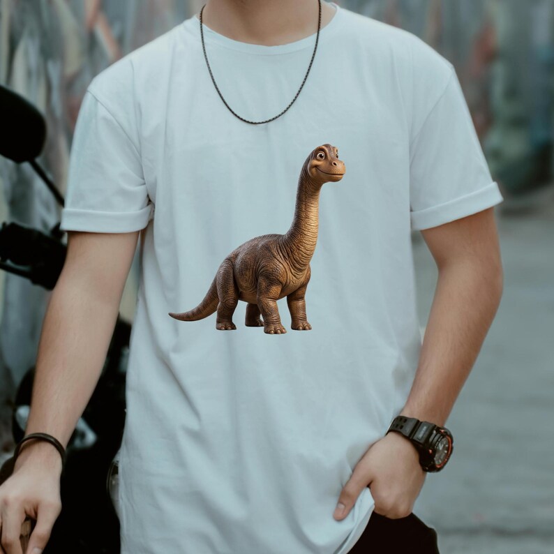 May include: White t-shirt featuring a brown dinosaur graphic. The dinosaur has a long neck and a friendly expression. The shirt is made of a soft, light-colored material and has short sleeves.