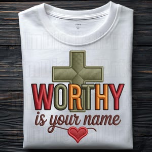 May include: White crew neck t-shirt with the embroidered text "WORTHY is your name" in red, orange, and brown. A green cross and a red heart are also embroidered on the shirt. The shirt is made of a soft, comfortable material.