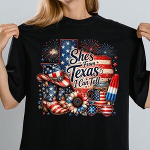 Pode incluir: Uma t-shirt preta com um gr&aacute;fico do estado do Texas com padr&otilde;es da bandeira americana, fogo de artif&iacute;cio, um chap&eacute;u de cowboy, botas, um girassol, um frasco, &oacute;culos em forma de cora&ccedil;&atilde;o e um gelado, com o texto She's From Texas I Can Tell.