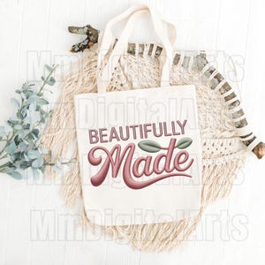 May include: A white tote bag with the words "BEAUTIFULLY Made" in a script font. The text is in a mauve color with a leaf design. The bag is on a white surface with decorative elements.