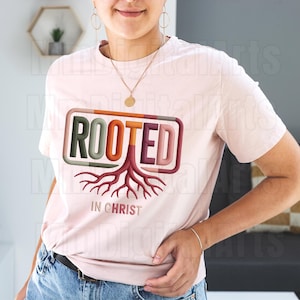 May include: A light pink t-shirt with the word "ROOTED" in a rectangular frame, featuring a tree root design. The text "IN CHRIST" is below the graphic. The shirt is a crew neck style.