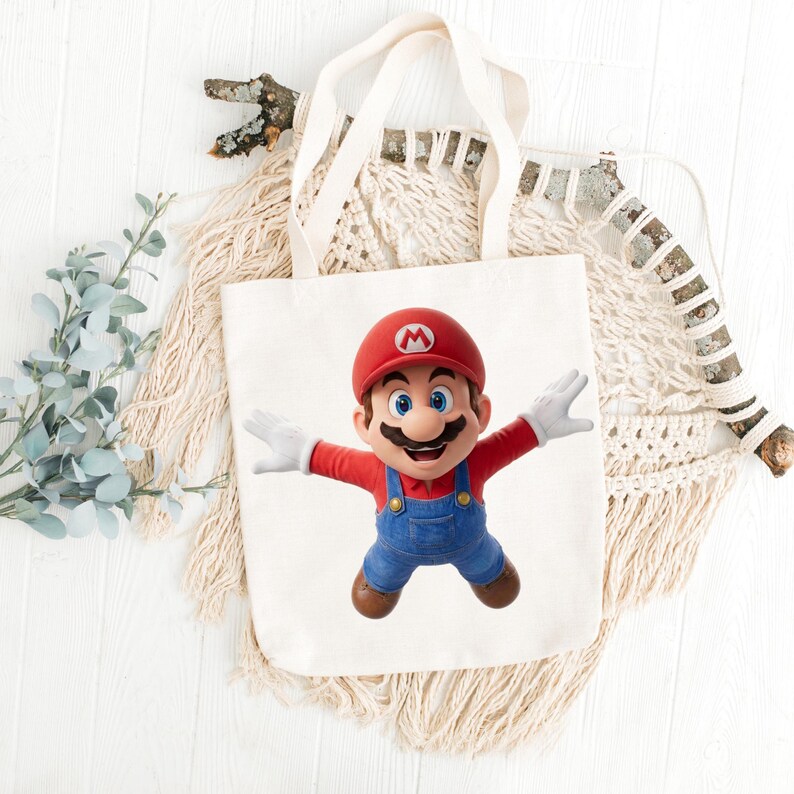 May include: A white canvas tote bag featuring a colorful image of Mario, the iconic video game character. Mario is depicted with his arms outstretched, wearing his signature red hat, blue overalls, and white gloves. The bag has two handles.