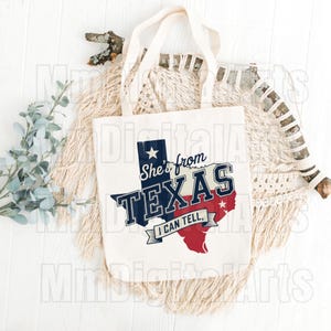 May include: Cream tote bag featuring the text "She's from TEXAS I CAN TELL" in navy blue, white, and red, with the outline of Texas. The bag has two cream-coloured handles. The design is printed on the front.