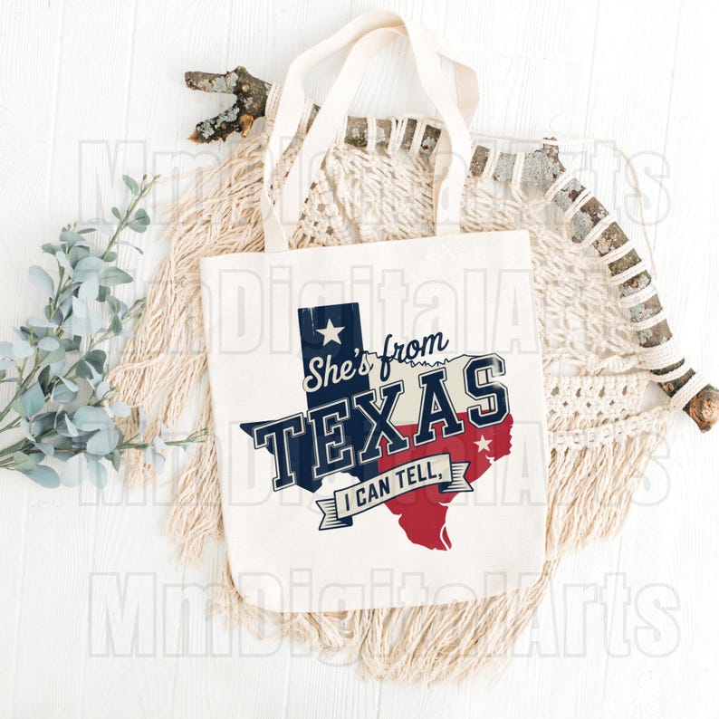 May include: Cream tote bag featuring the text "She's from TEXAS I CAN TELL" in navy blue, white, and red, with the outline of Texas. The bag has two cream-coloured handles. The design is printed on the front.