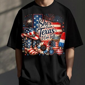 Pode incluir: Uma t-shirt preta com um gr&aacute;fico de uma bandeira em forma do Texas, um chap&eacute;u de cowboy, botas, um girassol, um frasco e um gelado, tudo com padr&otilde;es da bandeira americana. O texto "She's From Texas I Can Tell" est&aacute; rodeado por fogo de artif&iacute;cio.