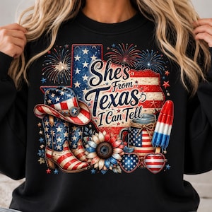 Pode incluir: Uma camisola preta com um gr&aacute;fico do estado do Texas com um padr&atilde;o de bandeira vermelha, branca e azul, fogo de artif&iacute;cio, um chap&eacute;u de cowboy, botas, um girassol, um frasco e um gelado. O texto diz: "She's From Texas I Can Tell."