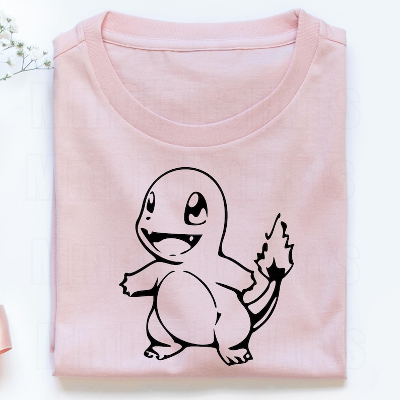 May include: A light pink t-shirt featuring a black graphic of a Charmander character. The design is centered on the front of the folded shirt. The shirt is made of a soft material.