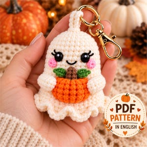 May include: A cream-colored crochet ghost keychain featuring a small orange pumpkin with green leaves, black safety eyes, and pink cheeks. The ghost is attached to a gold-tone metal lobster clasp. A circular badge displays the text PDF PATTERN IN ENGLISH.