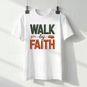 May include: White t-shirt with the text "WALK by FAITH" in green and orange block letters. The shirt is hanging on a hanger against a white background. The design includes a stylized footprint.