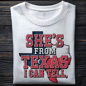 May include: White t-shirt featuring the text "SHE'S FROM TEXAS I CAN TELL," in red and blue. The design includes a Texas state outline in the same colours. The shirt is folded on a dark wood surface.