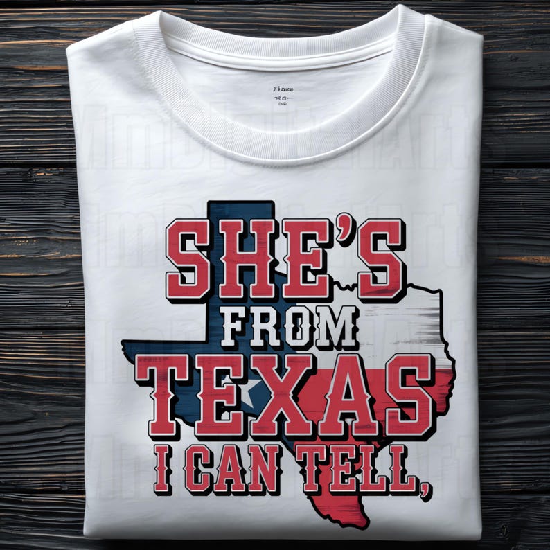 May include: White t-shirt featuring the text "SHE'S FROM TEXAS I CAN TELL," in red and blue. The design includes a Texas state outline in the same colours. The shirt is folded on a dark wood surface.