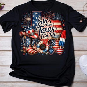 Pode incluir: Uma t-shirt preta com um gr&aacute;fico contendo o texto She's From Texas I Can Tell. O design inclui um chap&eacute;u de cowboy, uma bota, um girassol, um picol&eacute; e &oacute;culos em forma de cora&ccedil;&atilde;o, decorados com padr&otilde;es patri&oacute;ticos em vermelho, branco e azul.