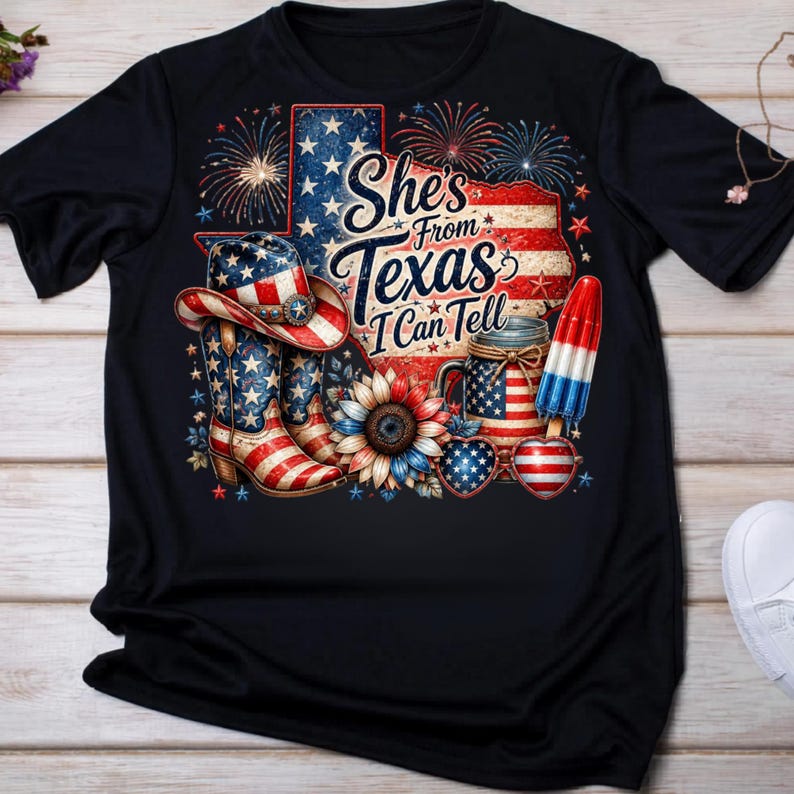 Pode incluir: Uma t-shirt preta com um gr&aacute;fico contendo o texto She's From Texas I Can Tell. O design inclui um chap&eacute;u de cowboy, uma bota, um girassol, um picol&eacute; e &oacute;culos em forma de cora&ccedil;&atilde;o, decorados com padr&otilde;es patri&oacute;ticos em vermelho, branco e azul.