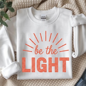 May include: White sweatshirt with the text "be the LIGHT" in orange. The design includes orange sun rays emanating from the words. The sweatshirt is folded neatly on a neutral-colored surface.