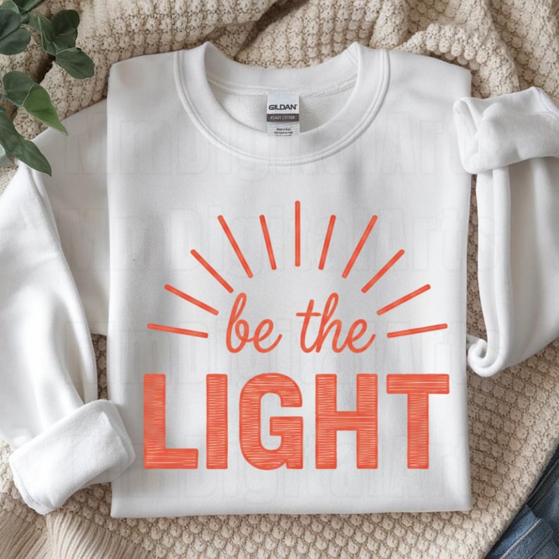May include: White sweatshirt with the text "be the LIGHT" in orange. The design includes orange sun rays emanating from the words. The sweatshirt is folded neatly on a neutral-colored surface.