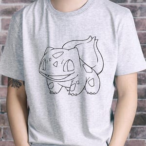 May include: A light gray t-shirt featuring a black and white outline of a Bulbasaur character. The Bulbasaur design is centered on the shirt and shows the character's distinctive features. The shirt is made of a soft material.