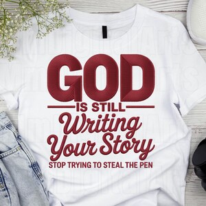 May include: White t-shirt with the embroidered text "GOD IS STILL Writing Your Story STOP TRYING TO STEAL THE PEN" in a burgundy color. The shirt is made of a soft material and is suitable for casual wear.