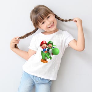 May include: White t-shirt featuring a colorful graphic of two cartoon characters riding a green dinosaur. The characters are wearing red and green hats. The t-shirt is worn by a young person with brown hair.