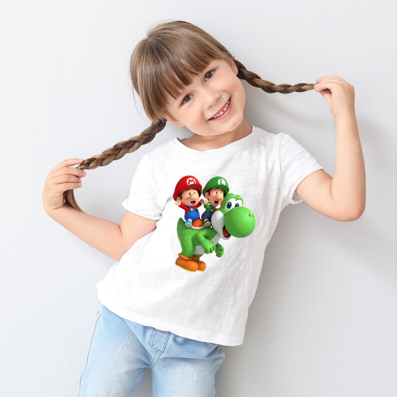 May include: White t-shirt featuring a colorful graphic of two cartoon characters riding a green dinosaur. The characters are wearing red and green hats. The t-shirt is worn by a young person with brown hair.
