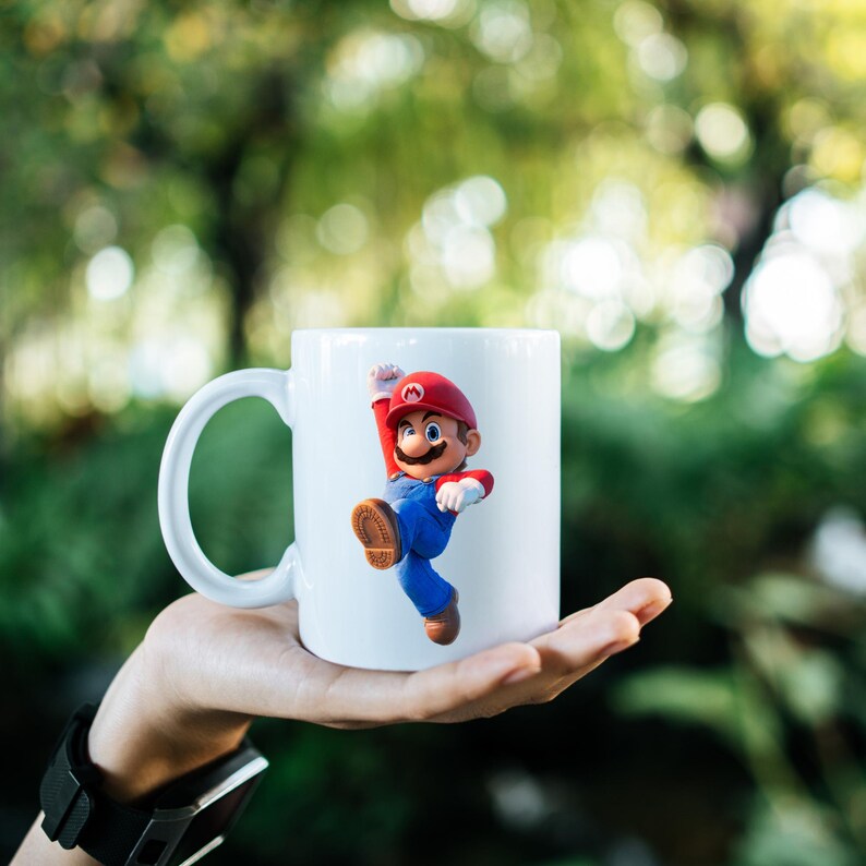 May include: White ceramic mug featuring a colorful Mario figure in a dynamic pose. The figure is wearing a red hat, blue overalls, and brown shoes. The mug has a large, curved handle.